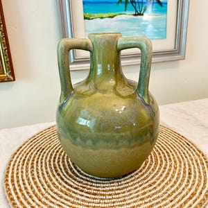 May include: A ceramic vase with a rounded body and two symmetrical handles extending from the neck to the shoulder. The piece features a glossy glaze in varying shades of olive green and sage, with a darker, mottled finish near the top rim.