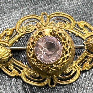Op de afbeelding: Vintage goudkleurige broche met een centrale, facetgeslepen, lichtpaarse edelsteen. De broche heeft een ovaal filigraan ontwerp met ingewikkeld krulwerk en een sluiting met speld. Decoratieve elementen zijn schelpachtige accenten.