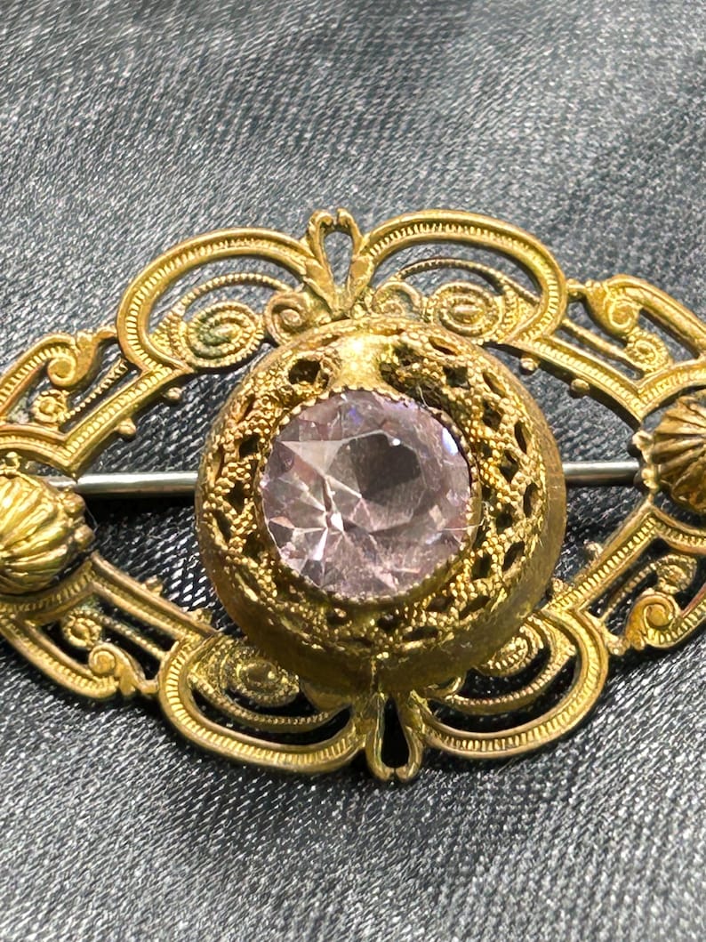 Op de afbeelding: Vintage goudkleurige broche met een centrale, facetgeslepen, lichtpaarse edelsteen. De broche heeft een ovaal filigraan ontwerp met ingewikkeld krulwerk en een sluiting met speld. Decoratieve elementen zijn schelpachtige accenten.
