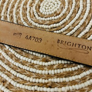 May include: Close-up of a brown leather belt with the brand name "BRIGHTON" stamped on it. The belt has the numbers "5/8" and "4A703" printed on it. The belt is lying on a woven, circular mat with white beads.