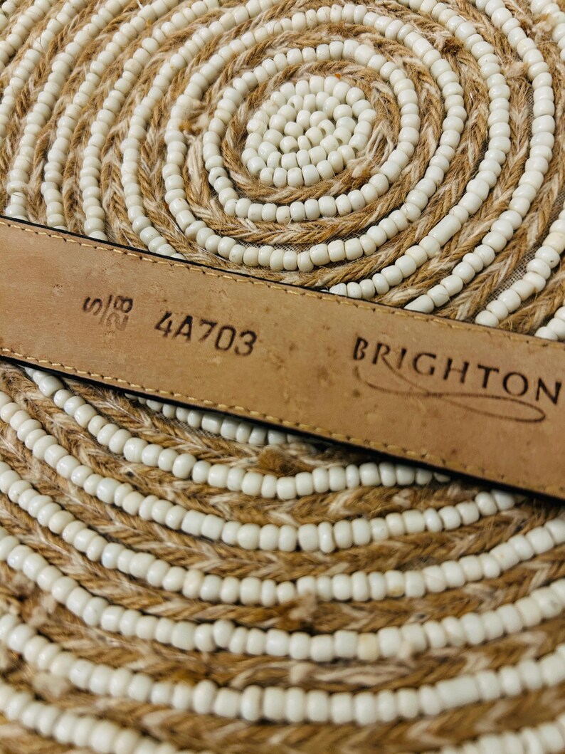 May include: Close-up of a brown leather belt with the brand name "BRIGHTON" stamped on it. The belt has the numbers "5/8" and "4A703" printed on it. The belt is lying on a woven, circular mat with white beads.