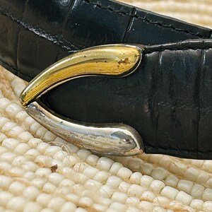 May include: Close-up of a black leather belt with a gold and silver-toned buckle. The belt has a textured, crocodile-like pattern. The buckle is curved and has a two-tone metallic finish.