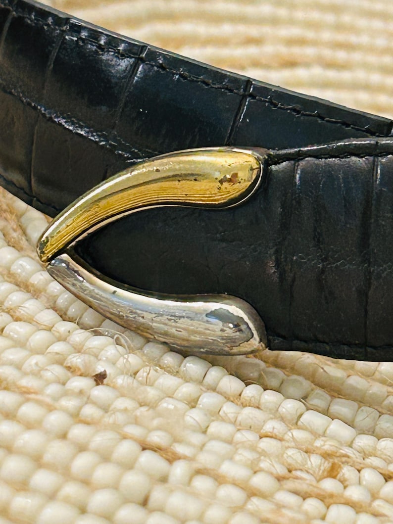 May include: Close-up of a black leather belt with a gold and silver-toned buckle. The belt has a textured, crocodile-like pattern. The buckle is curved and has a two-tone metallic finish.