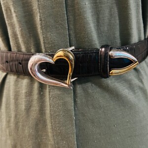 May include: Black belt with a silver and gold heart-shaped buckle. The belt has a textured, crocodile-like pattern. The buckle is a heart shape with a silver finish on the outer edges and a gold finish in the center.