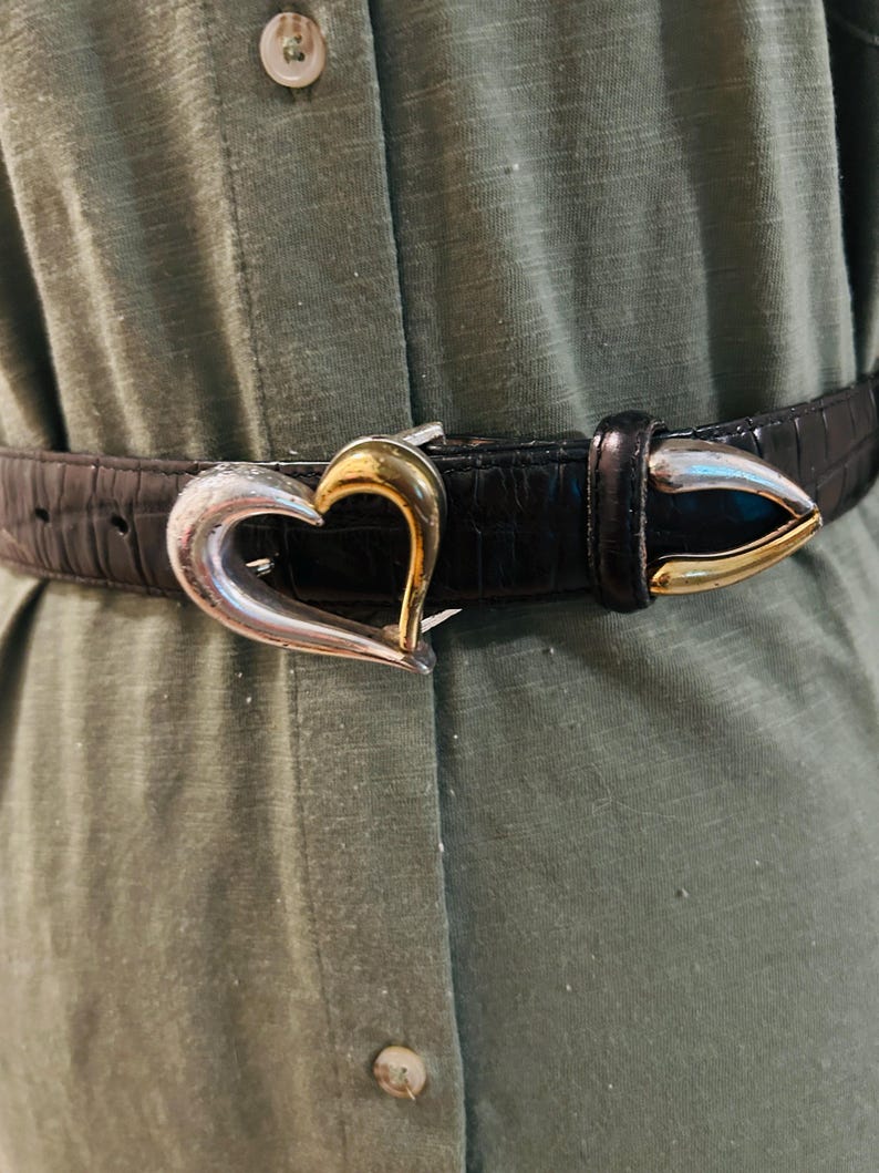 May include: Black belt with a silver and gold heart-shaped buckle. The belt has a textured, crocodile-like pattern. The buckle is a heart shape with a silver finish on the outer edges and a gold finish in the center.