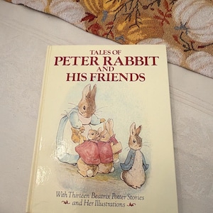 May include: A hardcover book titled Tales of Peter Rabbit and His Friends with Thirteen Beatrix Potter Stories and Her Illustrations. The cream-colored cover features a central illustration of four rabbits in clothing, including a blue jacket.