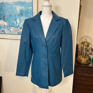 K&ouml;nnte beinhalten: T&uuml;rkisfarbener Lederblazer mit Reverskragen und Dreiknopfverschluss. Die Jacke wird auf einer wei&szlig;en Schaufensterpuppe pr&auml;sentiert. Der Blazer hat lange &Auml;rmel und eine taillierte Passform. Im Hintergrund h&auml;ngt ein Gem&auml;lde in Blau- und Wei&szlig;t&ouml;nen.