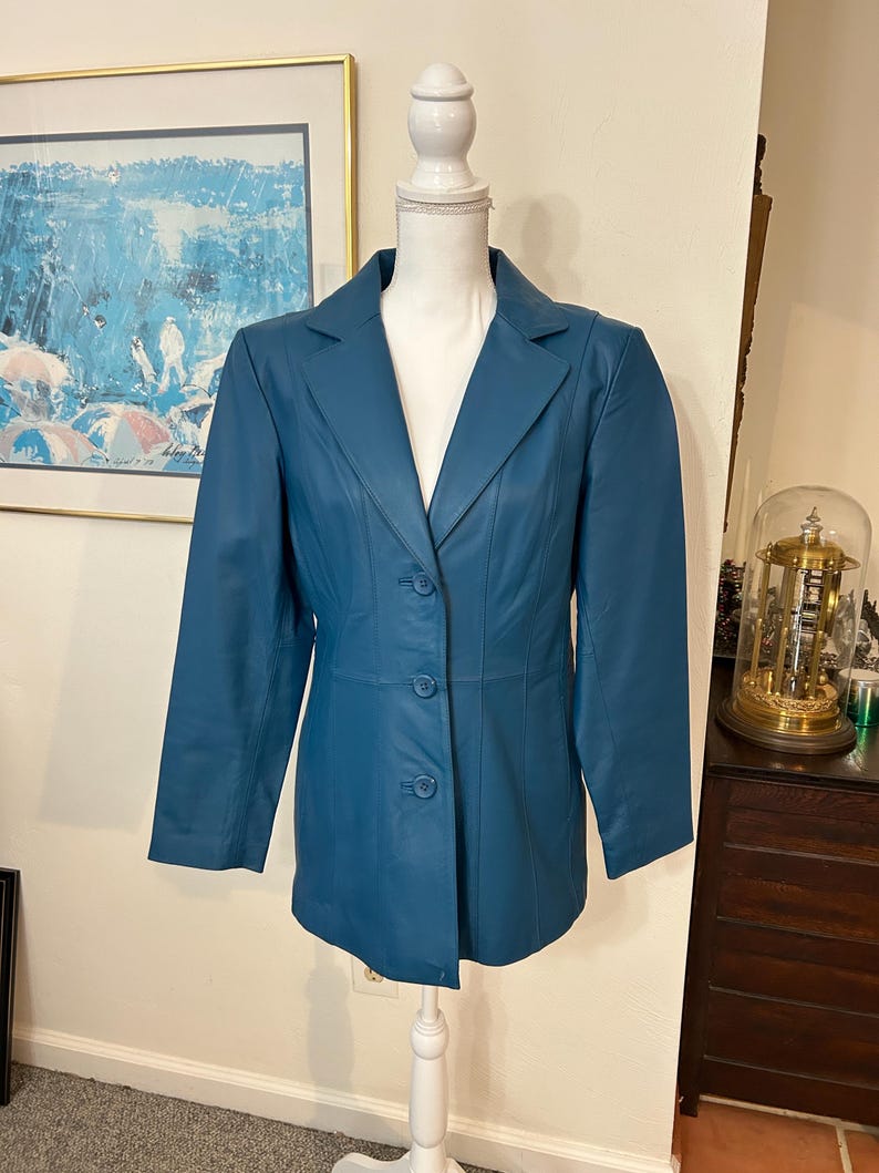 K&ouml;nnte beinhalten: T&uuml;rkisfarbener Lederblazer mit Reverskragen und Dreiknopfverschluss. Die Jacke wird auf einer wei&szlig;en Schaufensterpuppe pr&auml;sentiert. Der Blazer hat lange &Auml;rmel und eine taillierte Passform. Im Hintergrund h&auml;ngt ein Gem&auml;lde in Blau- und Wei&szlig;t&ouml;nen.