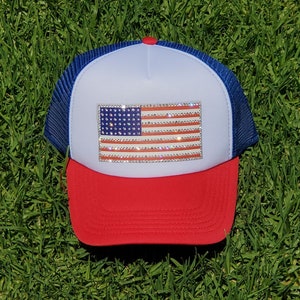 May include: A red, white, and blue trucker hat with a rhinestone American flag design. The hat has a blue mesh back and a white front panel.