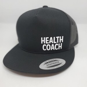 May include: Black and grey trucker hat with a white embroidered logo that says "HEALTH COACH". The hat has a black mesh back and a black snap closure.