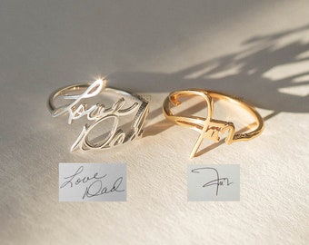 May include: Two rings, one silver and one gold, featuring handwritten script designs. The silver ring spells "Love Dad", and the gold ring has a signature-style design. Small cards below each ring display the same handwriting.