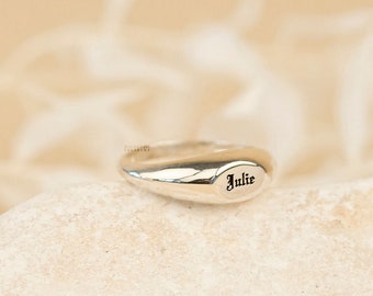 May include: A silver ring with the name "Julie" engraved on it. The ring is sitting on a light brown stone.