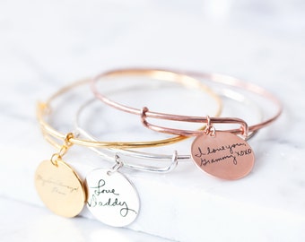Puede incluir: Tres pulseras de brazalete doradas con dijes. Los dijes son redondos y están grabados con mensajes escritos a mano. Los dijes dicen "Love Daddy", "I love you Grammy xoxo" y un mensaje ilegible.