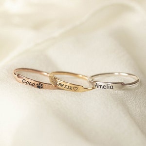 May include: Three thin gold rings with personalized engravings. The top ring says "Amelia", the middle ring says "MM XIX" with a heart symbol, and the bottom ring says "Coco" with a paw print.