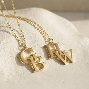 May include: Two gold-toned necklaces featuring delicate chains and pendant charms. One pendant displays the overlapping letters G and B, while the other pendant features the overlapping letters H and W in a serif font style.