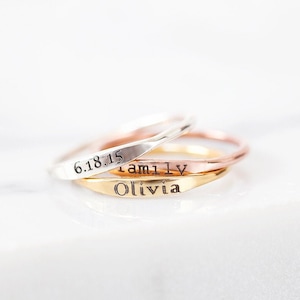 May include: Three thin silver, gold, and rose gold rings stacked on top of each other. The rings are engraved with the words "6.18.15", "Family", and "Olivia".