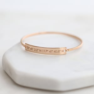 May include: A gold-toned bangle bracelet with a rectangular tag engraved with the coordinates "33° 41' 2.2" N 117° 47' 40.8" W".