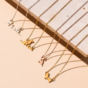 May include: Five gold necklaces with different animal pendants. The pendants are in the shape of a cat, a dog, and a dachshund. Each pendant has a letter engraved on it. The necklaces are on a light brown surface.