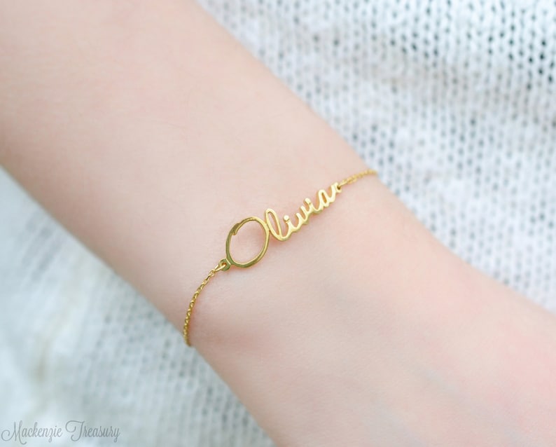 May include: Gold bracelet featuring the name "Olivia" in a cursive script. The bracelet includes a small circular charm.