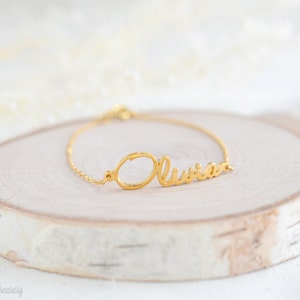 May include: Gold name bracelet featuring the name "Olivia" in a cursive script. The bracelet is displayed on a light wood surface.