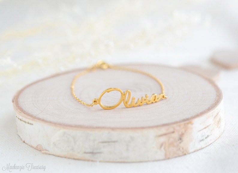 May include: Gold name bracelet featuring the name "Olivia" in a cursive script. The bracelet is displayed on a light wood surface.