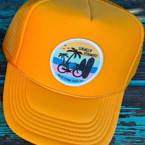May include: A bright yellow trucker hat with a mesh back and a curved brim. The hat features a circular patch with the words "Locally Stranded" above a design of palm trees, a bicycle, and a surfboard. The text "Silver Strand Beach, CA" is below the design.