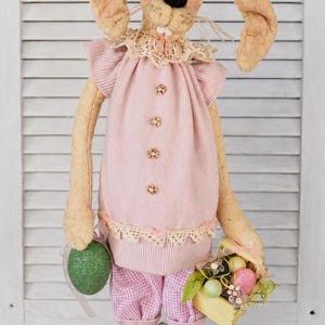 May include: A plush tan rabbit doll with long ears, wearing a pink ribbed top with flower-shaped buttons, a lace collar, and pink gingham pants. The rabbit holds a green glittery egg and a small yellow basket filled with colorful miniature eggs.