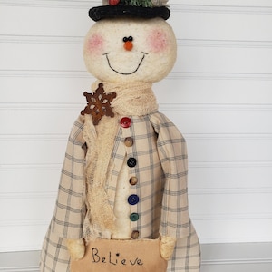 May include: A handmade snowman doll wearing a plaid coat, a scarf, and a black top hat with a sprig of greenery and red berries. The snowman is holding a sign that says "Believe".