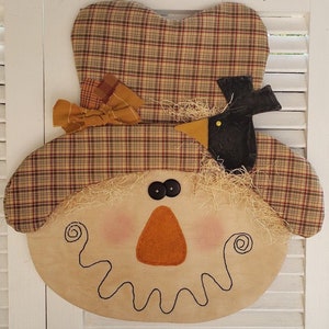 May include: A handmade scarecrow face with a plaid hat, a black bird, and a straw beard. The face is made of burlap and has a stitched smile, orange nose, and black eyes.