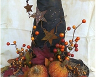 May include: A black witch's hat with a pointed top and a wide brim, decorated with fall foliage, berries, and small stars. The hat is sitting on a bed of moss and twigs.