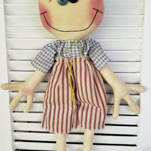 May include: A handmade rag doll with brown hair, a blue and white checkered shirt, red and white striped pants, and black shoes. The doll has a smiling face with rosy cheeks and blue eyes.
