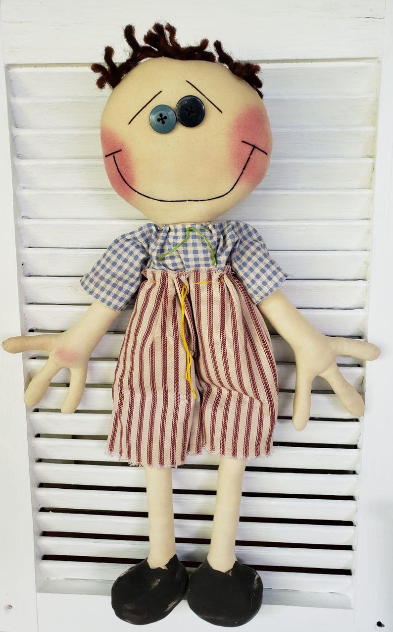 May include: A handmade rag doll with brown hair, a blue and white checkered shirt, red and white striped pants, and black shoes. The doll has a smiling face with rosy cheeks and blue eyes.