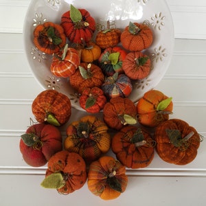 May include: A collection of small, handmade fabric pumpkins in various shades of orange and brown. The pumpkins are arranged in a white bowl and on a white surface.