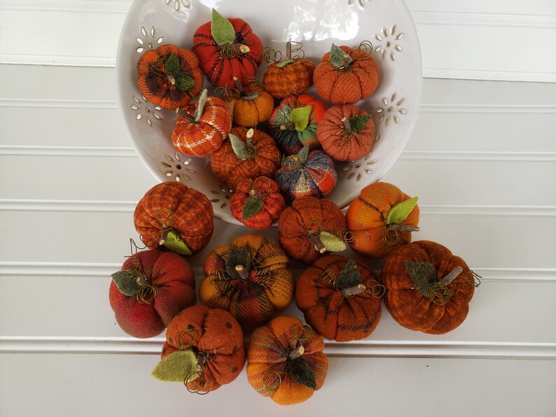 May include: A collection of small, handmade fabric pumpkins in various shades of orange and brown. The pumpkins are arranged in a white bowl and on a white surface.