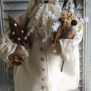 May include: A white felt Santa Claus doll with a white beard and a white hat. The doll is holding a small brown tree and a bundle of twigs with white snowflakes. The doll is standing on a wooden base with white snow.