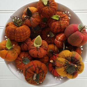 May include: A white bowl filled with handmade fabric pumpkins in various shades of orange, red, and brown. The pumpkins have a rustic, homespun look and are perfect for fall decor.