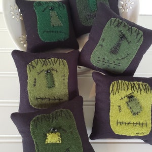 May include: Six small black felt pillows with green felt Frankenstein faces. Each pillow has black bead eyes and stitched details.