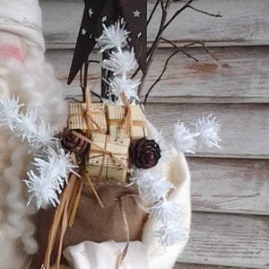 May include: A white, fuzzy, snowman-like figure with a brown sack filled with wrapped gifts and a rustic star on top. The figure is decorated with white, fluffy, snowflake-like embellishments and brown twigs.