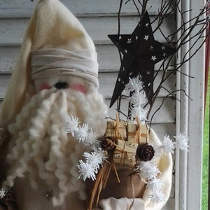 May include: A white felt Santa Claus doll with a long white beard and a white hat. The doll is holding a burlap sack filled with small gifts and a star-shaped ornament. The doll is standing in front of a white wooden wall.