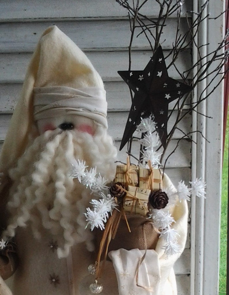 May include: A white felt Santa Claus doll with a long white beard and a white hat. The doll is holding a burlap sack filled with small gifts and a star-shaped ornament. The doll is standing in front of a white wooden wall.