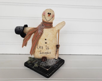 May include: A handmade snowman figurine with a black top hat, striped scarf, and a broom. The snowman is standing on a wooden base with the words "Let it Snow" written on it.