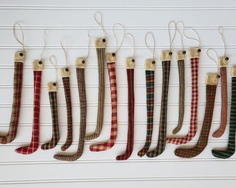 May include: A collection of 16 handmade fabric hockey stick ornaments in various plaid and striped patterns. The ornaments are approximately 6 inches long and feature a brown button at the top for hanging.