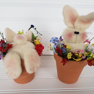 May include: Two white bunny figurines are sitting in terracotta flower pots filled with colorful artificial flowers. The bunnies are made of soft, fluffy material and have pink noses and black eyes.