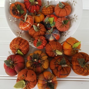 May include: A collection of handmade fabric pumpkins in various shades of orange, red, and brown. The pumpkins are arranged in a white bowl and on a white surface.
