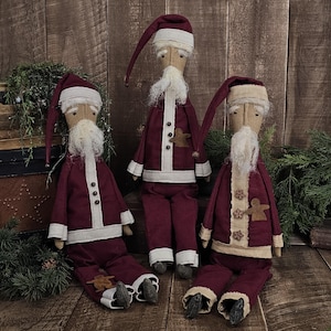 May include: Three fabric Santa Claus dolls in deep burgundy outfits with cream-colored trim, long pointed hats, and white yarn beards. Each doll features small brown gingerbread man accents on the clothing and dark grey fabric shoes.