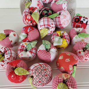May include: A collection of handmade patchwork fabric apples in red, white, and green. The apples are arranged in a bowl and on a white surface.