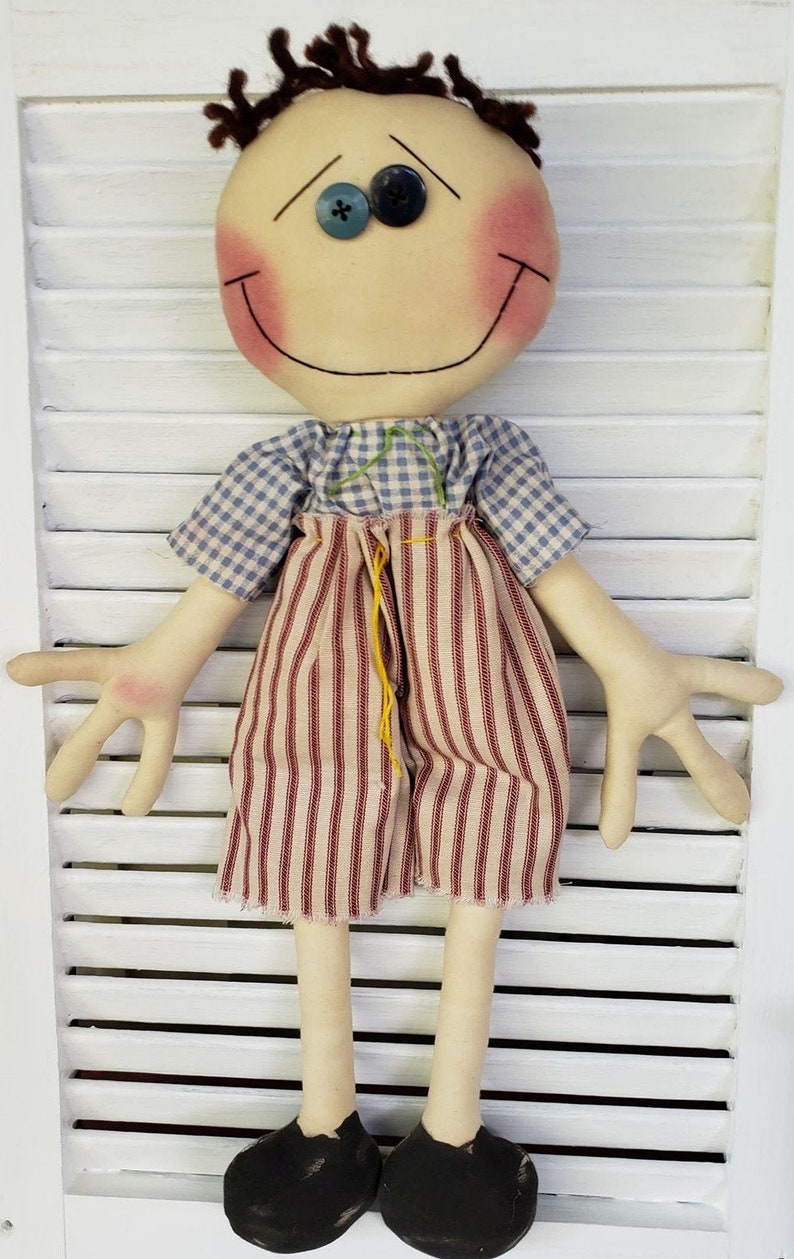 May include: A handmade rag doll with a smiling face, blue eyes, and rosy cheeks. The doll is wearing a blue and white checkered shirt, red and white striped pants, and black shoes.