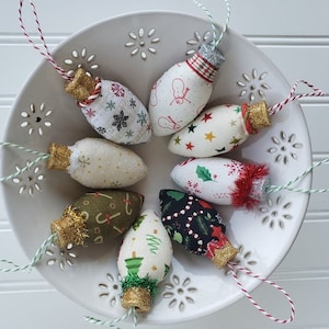 May include: Eight handmade fabric Christmas decorations shaped like light bulbs. Each ornament is decorated with a different festive pattern and has a gold glitter top and a white string for hanging.