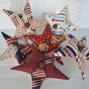 Könnte beinhalten: Fünf rote, weiße und blaue Stoffsterne mit einer Glocke an jedem. Die Sterne sind aus Patchwork-Stoff mit verschiedenen patriotischen Mustern.