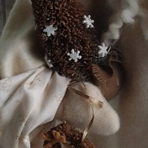 May include: A white fabric Santa Claus figure with a brown, twig-like Christmas tree in its hand. The tree is decorated with white snowflake ornaments.